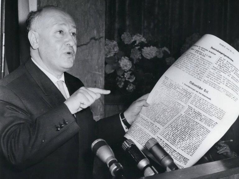 Ein Mann Mitte fünfzig mit Glatze hält ein Dokument in der linken Hand und zeigt mit dem Zeigefinger der rechten auf das Dokument.