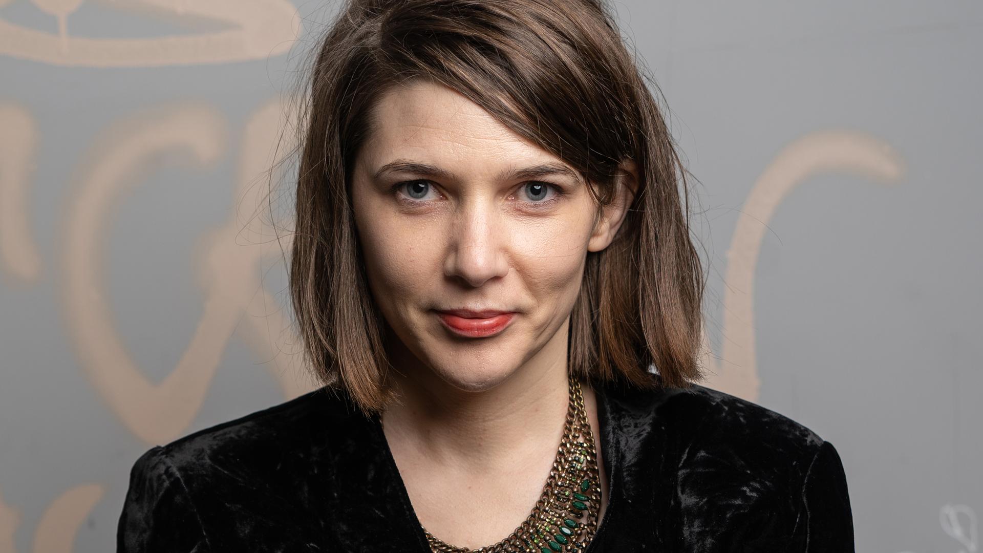 Vor grauem Hintergrund mit Graffiti schaut Anaïs Meier in schwarzem Samtblazer in die Kamera. Vor grauem Hintergrund mit Graffiti schaut Anaïs Meier in schwarzem Samtblazer in die Kamera.