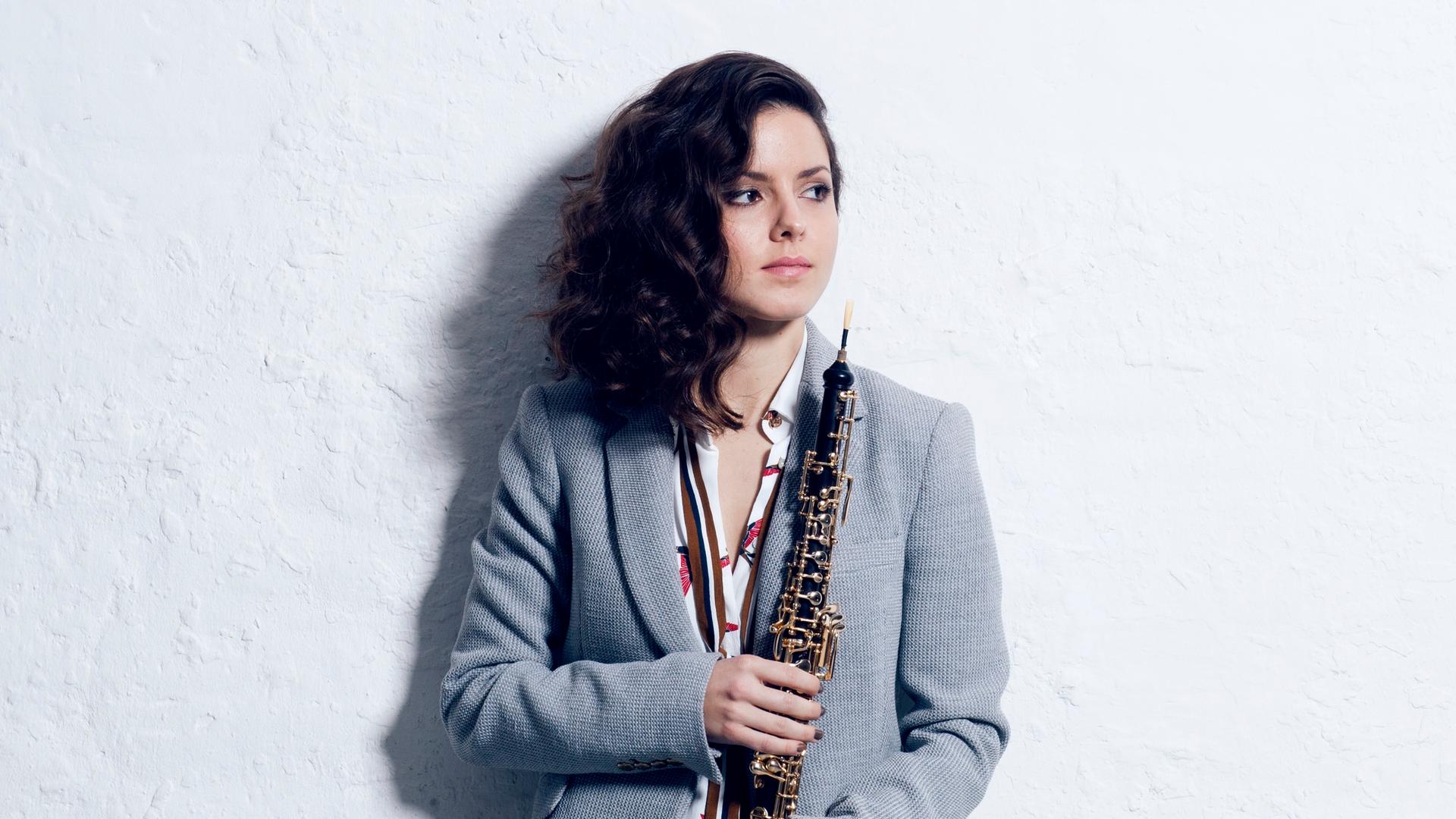Cristina Gómez Godoy im Anzug steht mit langen, lockigen, offenen Haaren an einer Wand angeleht, blickt zur Seite und hält ihre Oboe umarmt vor sich.