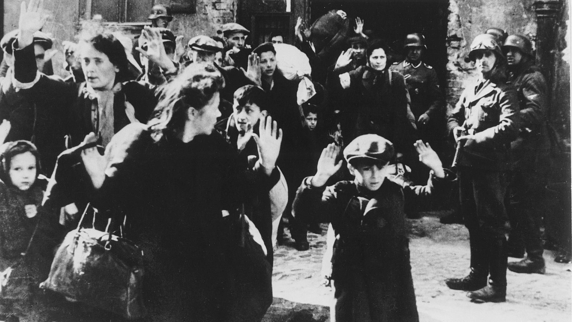 Historisches Foto vom Abtransport von Juden aus dem Warschauer Ghetto im Jahr 1943. Frauen und Kinder gehen mit erhobenen Händen aus einem Haus, Soldaten stehen mit Waffen daneben. Vorne Dab Siemiatek oder Zvi Nussbaum, oder Levi Zelinwarger, daneben seine Mutter Chana Zelinwarger, (l.) Hanka Lamet, (2.v.li.) M. Lamet Goldfinger, Leo Kartuzinski (mit Bündel), (r.) Golda Stavarowski und (2.v.re.) SS-Mann Josef Blösche.
