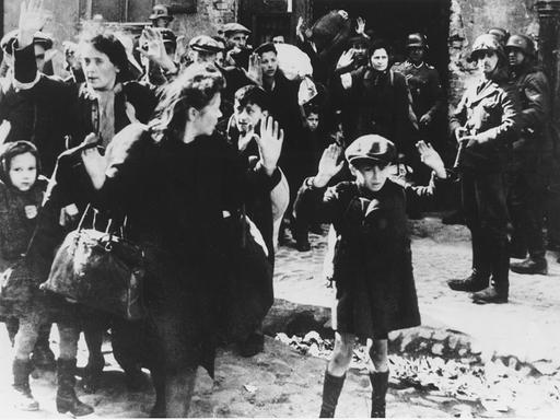 Historisches Foto vom Abtransport von Juden aus dem Warschauer Ghetto im Jahr 1943. Frauen und Kinder gehen mit erhobenen Händen aus einem Haus, Soldaten stehen mit Waffen daneben. Vorne Dab Siemiatek oder Zvi Nussbaum, oder Levi Zelinwarger, daneben seine Mutter Chana Zelinwarger, (l.) Hanka Lamet, (2.v.li.) M. Lamet Goldfinger, Leo Kartuzinski (mit Bündel), (r.) Golda Stavarowski und (2.v.re.) SS-Mann Josef Blösche.