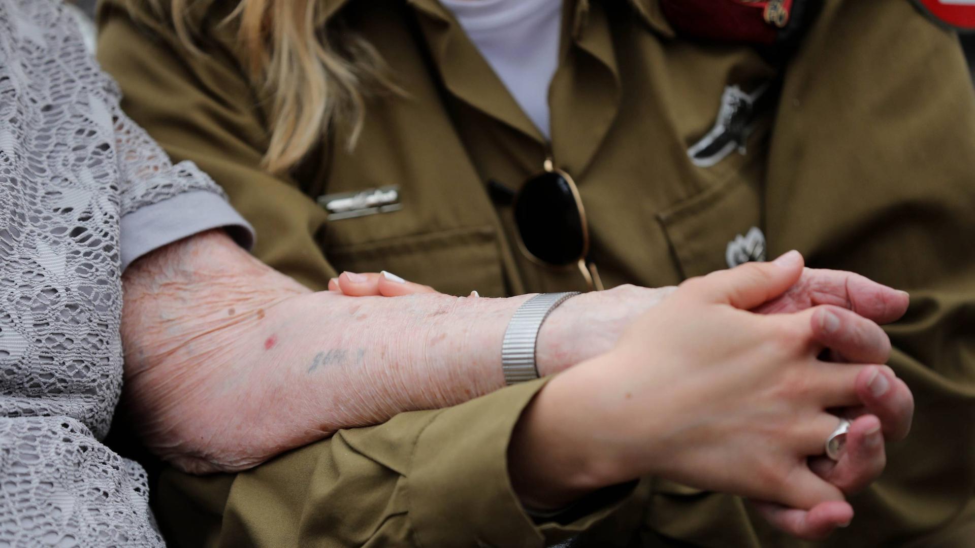 Holocaust-Überlebende Hanna Tessler mit ihrer Enkelin Shira Tessler. Eine israelische Soldatin hält die Hand ihrer Großmutter, die den Holocaust überlebt hat. Auf den Arm hat sie eine tätowierte Nummer. Holocaust-Überlebende Hanna Tessler mit ihrer Enkelin Shira Tessler. Eine israelische Soldatin hält die Hand ihrer Großmutter, die den Holocaust überlebt hat. Auf den Arm hat sie eine tätowierte Nummer.