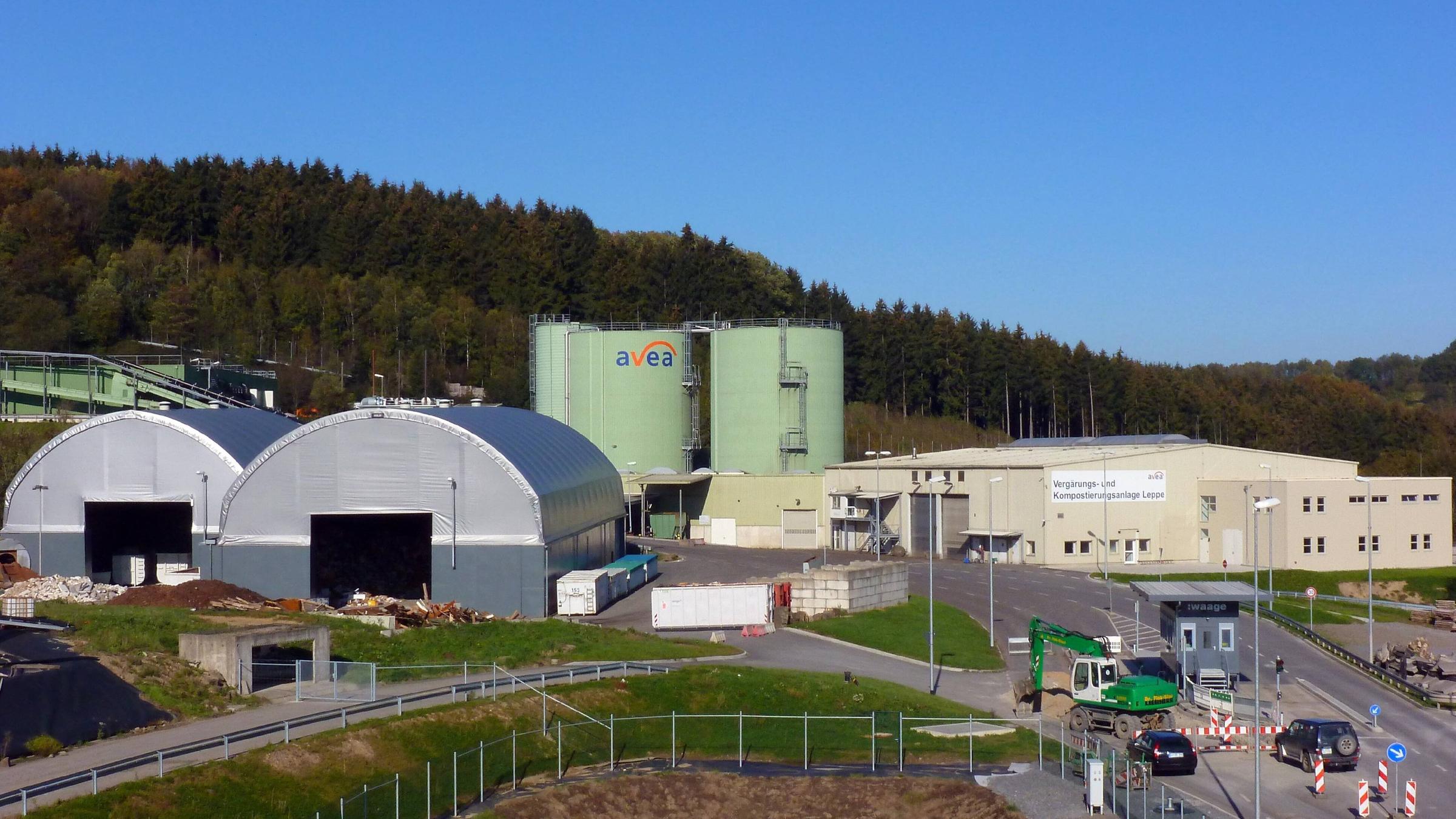 Zwei silberne Lagerhallen und dahinter zwei hellgrüne Silos stehen auf dem Gelände einer Mülldeponie vor einem Wald. Zwei silberne Lagerhallen und dahinter zwei hellgrüne Silos stehen auf dem Gelände einer Mülldeponie vor einem Wald.