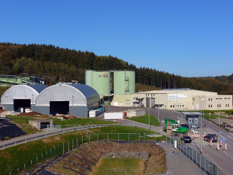 Zwei silberne Lagerhallen und dahinter zwei hellgrüne Silos stehen auf dem Gelände einer Mülldeponie vor einem Wald.