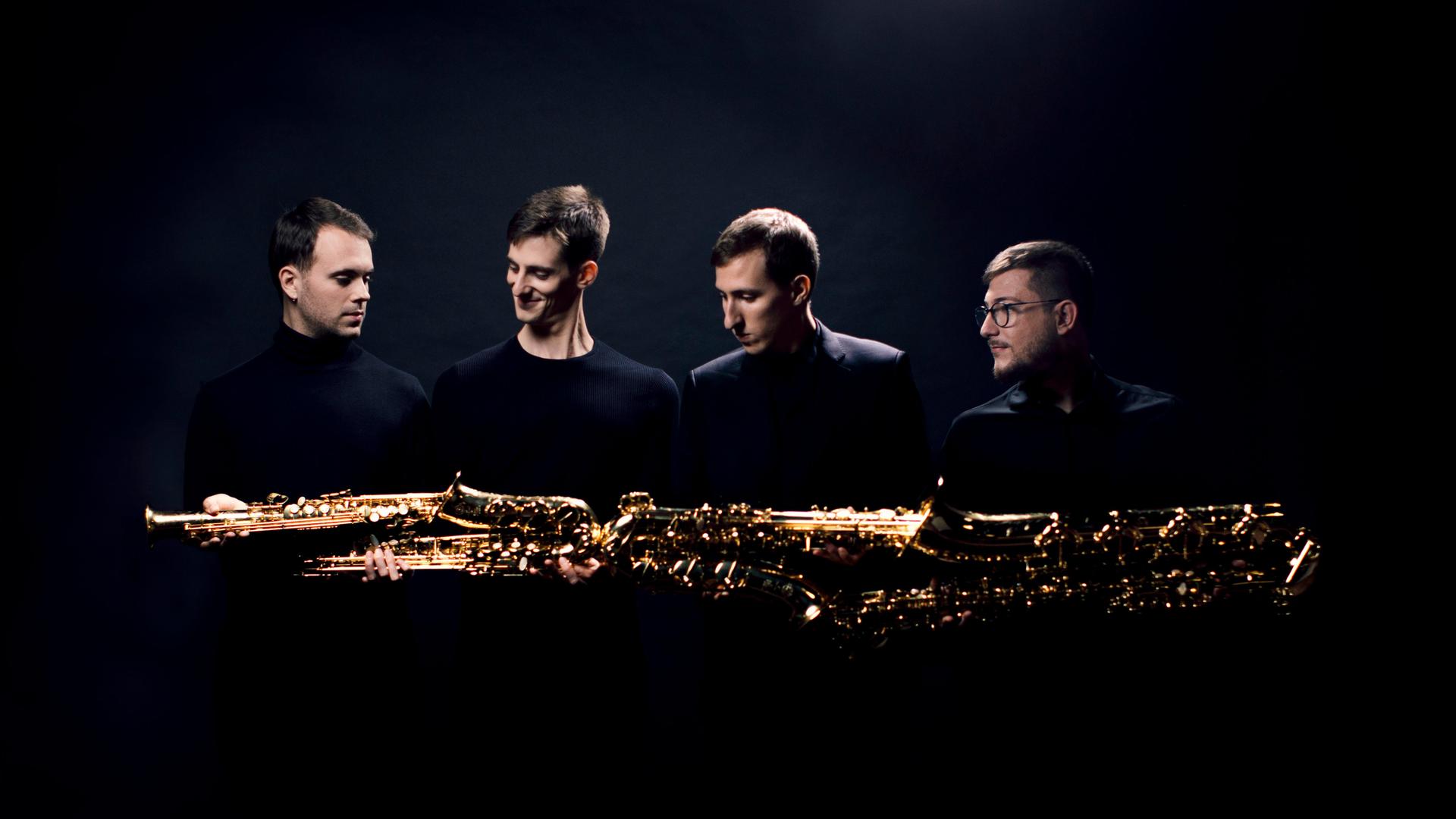Vier junge Männer halten ihre unterschiedlich großen Saxophone quer, sodass er schient, als ob die Saxophone ein riesiges, goldenes Instrument ergeben. Vier junge Männer halten ihre unterschiedlich großen Saxophone quer, sodass er schient, als ob die Saxophone ein riesiges, goldenes Instrument ergeben.