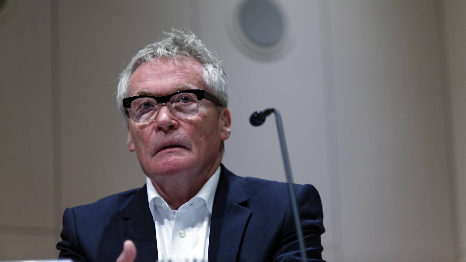Ein älterer Mann mit ergrautem Haar und Brille, gekleidet in einem dunkelblauen Anzug, sitzt an einem Mikrofon. Es handelt sich um den Architekten Arno Lederer. Ein älterer Mann mit ergrautem Haar und Brille, gekleidet in einem dunkelblauen Anzug, sitzt an einem Mikrofon. Es handelt sich um den Architekten Arno Lederer.