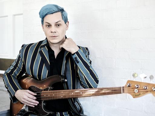 Jack White ist von der Hüfte an aufwärts zu sehen. Er hat die Haare hellblau gefärbt, sein Sakko ist in hellblau und  schwarz länggestreift, darunter trägt ein in dem hellen Raum ein schwarzes Oberteil. Vor dem Bauch hält er eine Bassgitarre.