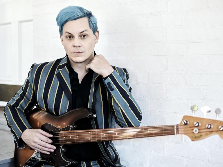 Jack White ist von der Hüfte an aufwärts zu sehen. Er hat die Haare hellblau gefärbt, sein Sakko ist in hellblau und  schwarz länggestreift, darunter trägt ein in dem hellen Raum ein schwarzes Oberteil. Vor dem Bauch hält er eine Bassgitarre.