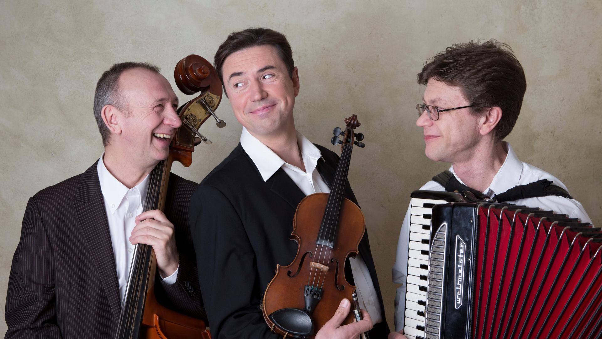 Drei Musiker sind zu sehen, sie lachen und halten ihre Instrumente fest. 