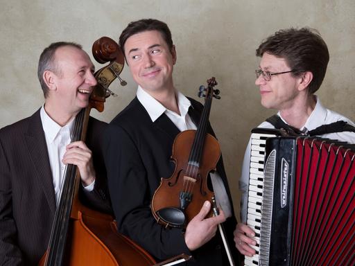 Drei Musiker sind zu sehen, sie lachen und halten ihre Instrumente fest. 