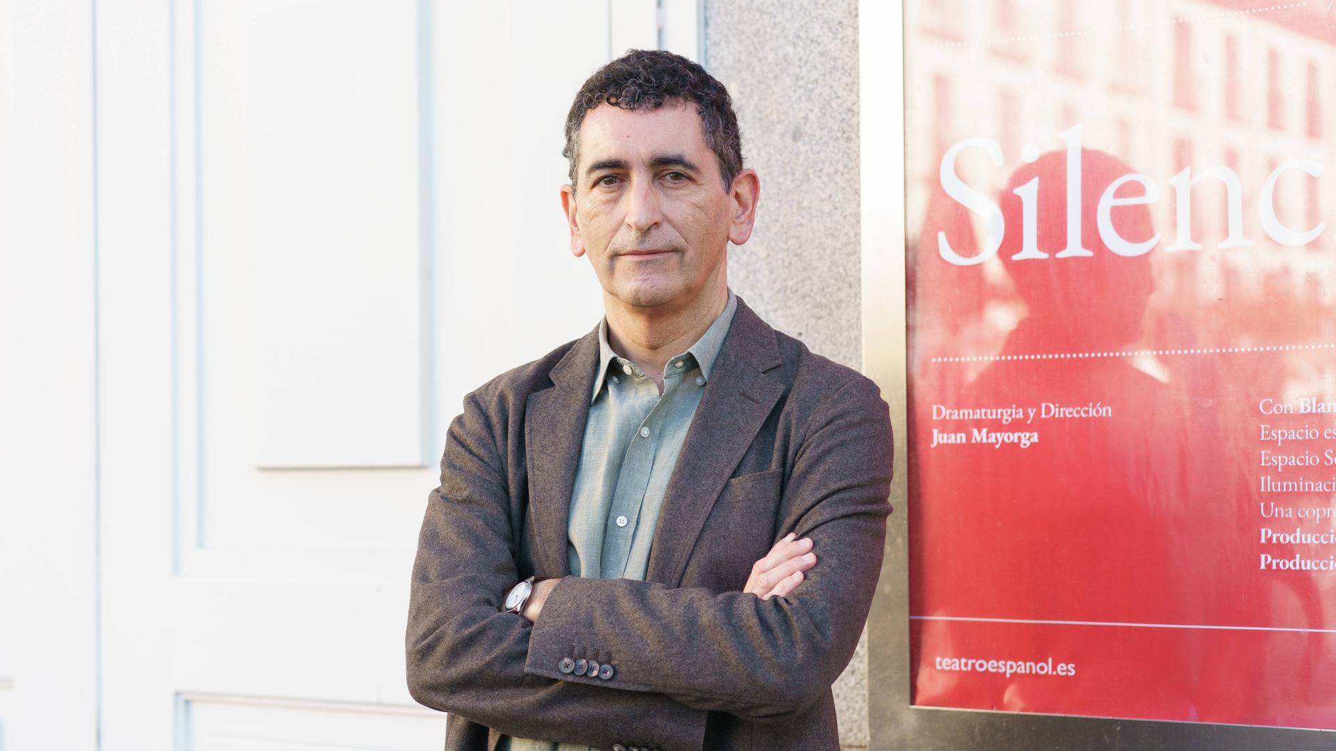 Der Autor Juan Mayorga steht mit verschränkten Armen vor einem Theaterplakat Der Autor Juan Mayorga steht mit verschränkten Armen vor einem Theaterplakat