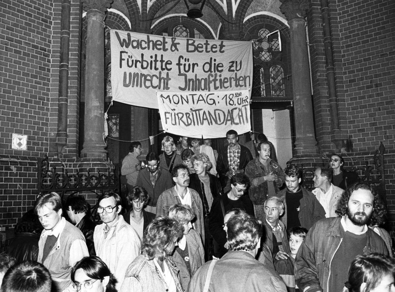 Schwarz-weiß Fotografie einer Menschengruppe, die sich am Abend vor dem Eingang einer Backsteinkirche versammelt hat.