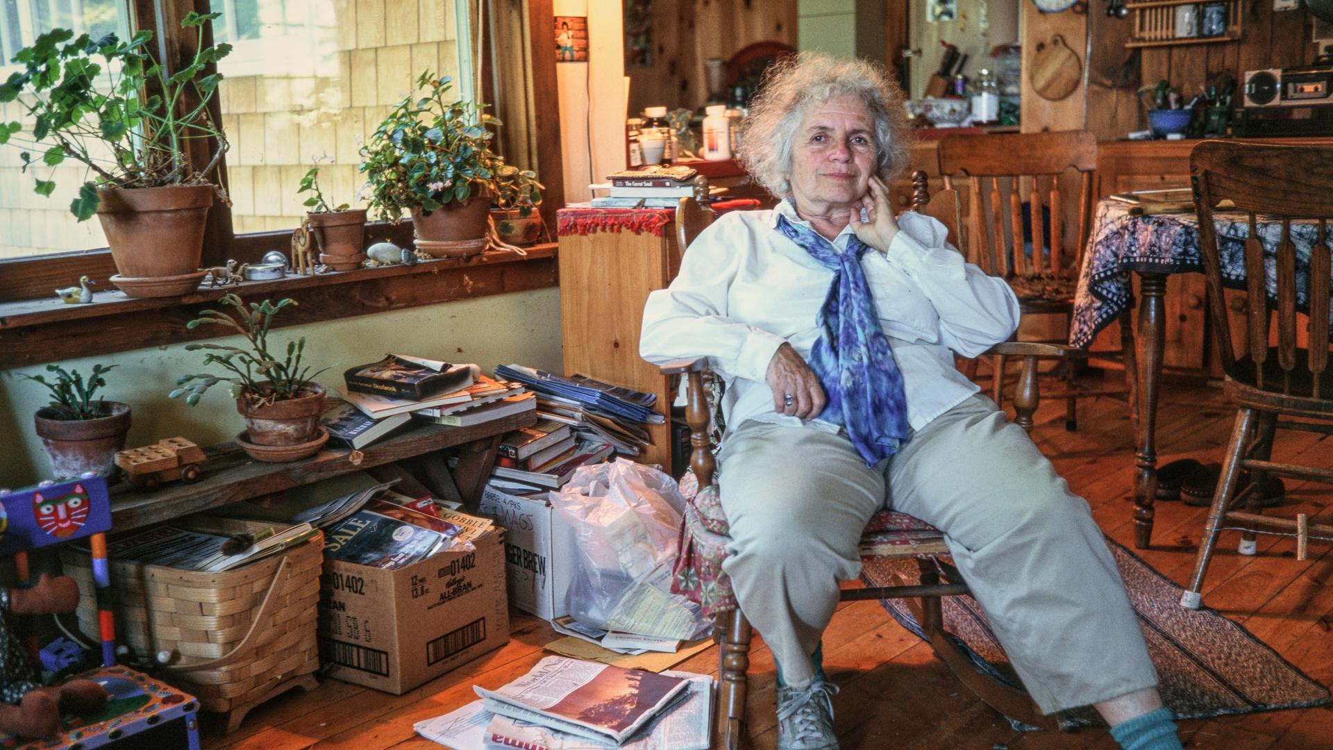 Grace Paley, eine ältere Frau mit grauen Locken, dreckigen Sneakers und einem blauen Halstuch, sitzt in ihrem Zuhause in einem hölzernen Schaukelstuhl, umgeben von Büchern, Pflanzen und Kartons voller Zeitschriften. Grace Paley, eine ältere Frau mit grauen Locken, dreckigen Sneakers und einem blauen Halstuch, sitzt in ihrem Zuhause in einem hölzernen Schaukelstuhl, umgeben von Büchern, Pflanzen und Kartons voller Zeitschriften.