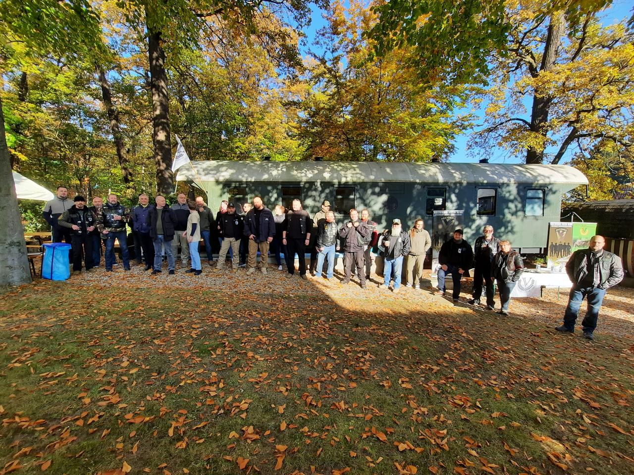 Vor einem umgebauten Bahnwaggon steht eine Gruppe aus Menschen, die meisten sind Männer, ein paar wenige Frauen sind auch dabei. Vor einem umgebauten Bahnwaggon steht eine Gruppe aus Menschen, die meisten sind Männer, ein paar wenige Frauen sind auch dabei.