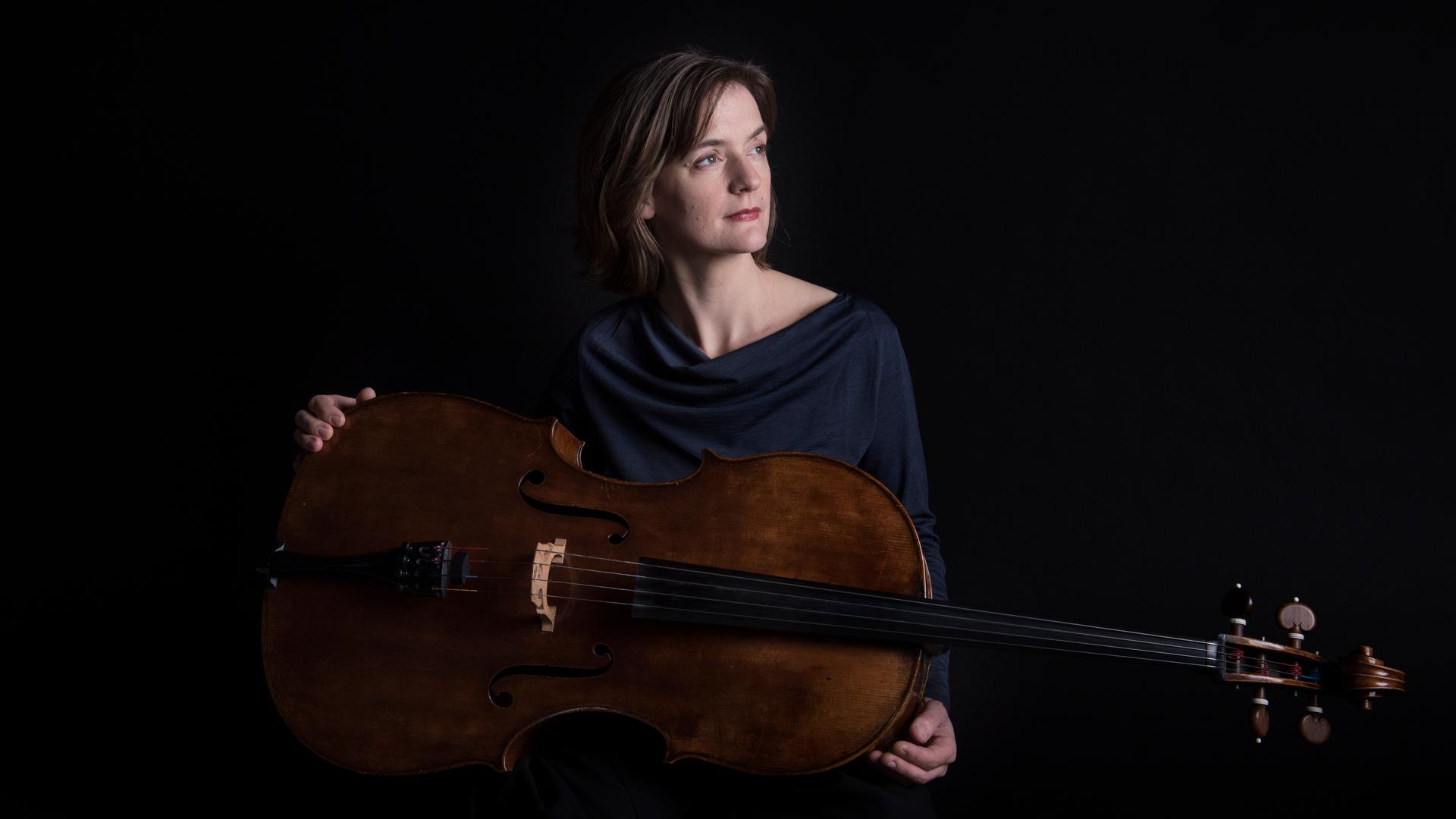 Eine Frau sitzt in dunkler Kleidung vor einem schwarzen Hintergrund. Sie blickt nachdenklich zur linken Seite. Über ihre Beine liegt quer ein Violoncello, das sie mit ihren Händen locker umfasst. Eine Frau sitzt in dunkler Kleidung vor einem schwarzen Hintergrund. Sie blickt nachdenklich zur linken Seite. Über ihre Beine liegt quer ein Violoncello, das sie mit ihren Händen locker umfasst.