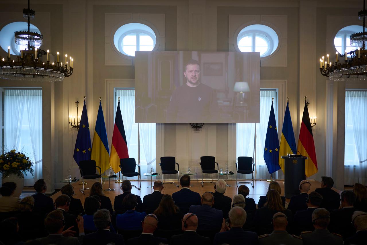 Der ukrainische Präsident Selenskyj ist auf einer Videoleinwand zu sehen. Im Saal sitzen zahlreiche Personen und hören ihm zu. Auf der Bühne stehen Stühle und Flaggen der EU, der Ukraine und der Bundesrepublik.