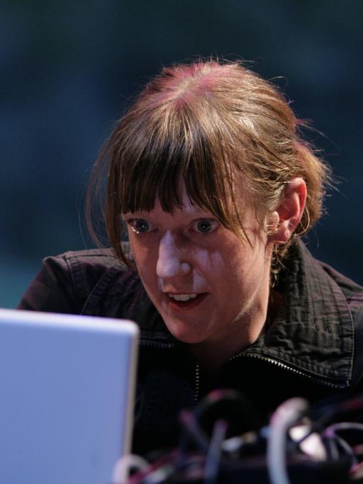 Eine junge Frau sitzt hinter einem Laptop und einem Mikrophon und schaut in den Bildschirm.