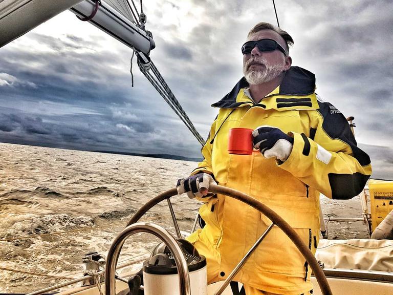 Torsten Mohr trägt Handschuhe, eine Sonnenbrille und eine gelbe Jacke. Er steht am Ruder seines Segelbootes. In der Hand hält er eine rote Tasse. Am Himmel sind graue Wolken.