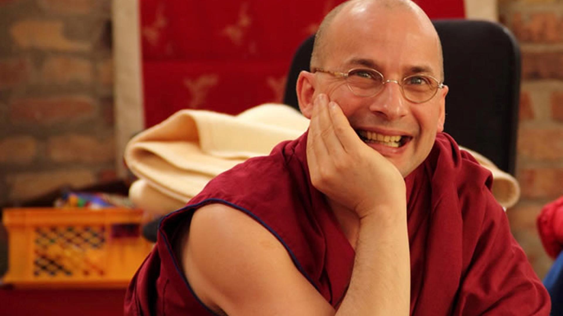 Tenzin Peljor stützt seinen Kopf auf einen Arm und lächelt in die Kamera. Er trägt eine rotes, kurzärmeliges Gewand. Tenzin Peljor stützt seinen Kopf auf einen Arm und lächelt in die Kamera. Er trägt eine rotes, kurzärmeliges Gewand.