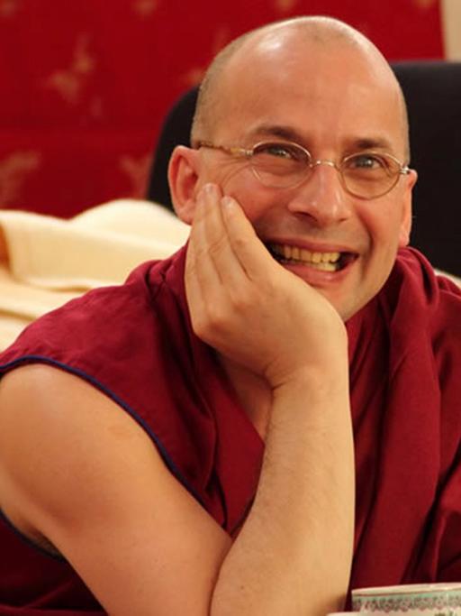 Tenzin Peljor stützt seinen Kopf auf einen Arm und lächelt in die Kamera. Er trägt eine rotes, kurzärmeliges Gewand.