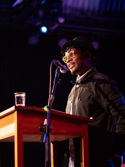 Der Autor, Fotograf und Filmemacher Lidudumalingani aus Johannesburg steht an einem Rednerpult. Er trägt Brille und Hut. 