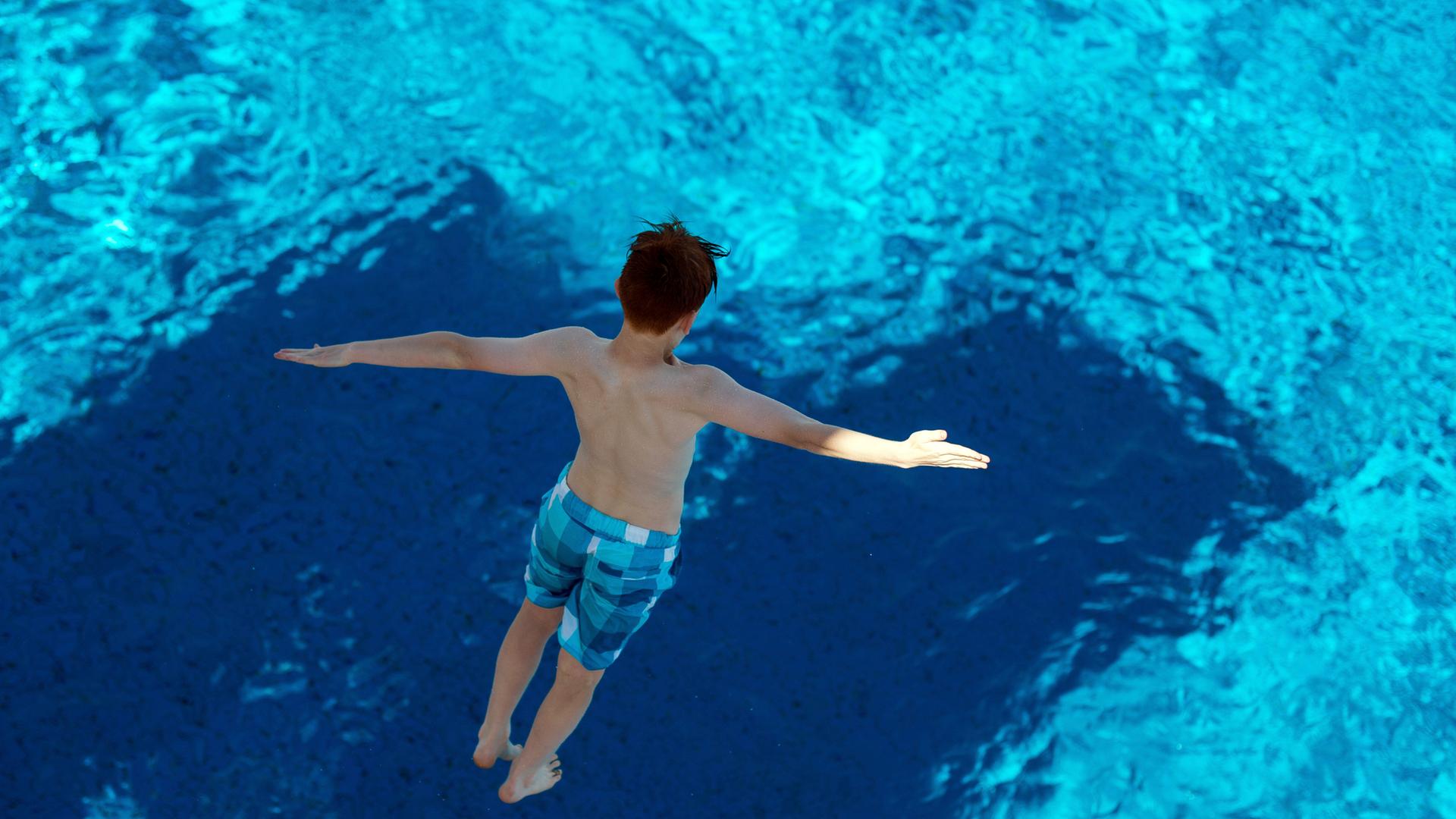 Ein Junge, in blau karierter Badehose und mit zur Seite ausgebreiteten Armen, springt von einem Sprungturm ins Wasser. Die Aufnahme zeigt ihn von oben, in der Luft schwebend. Ein Junge, in blau karierter Badehose und mit zur Seite ausgebreiteten Armen, springt von einem Sprungturm ins Wasser. Die Aufnahme zeigt ihn von oben, in der Luft schwebend.