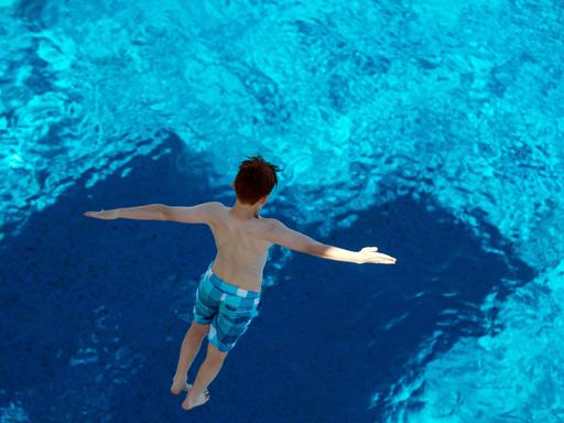 Ein Junge, in blau karierter Badehose und mit zur Seite ausgebreiteten Armen, springt von einem Sprungturm ins Wasser. Die Aufnahme zeigt ihn von oben, in der Luft schwebend.
