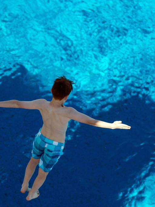 Ein Junge, in blau karierter Badehose und mit zur Seite ausgebreiteten Armen, springt von einem Sprungturm ins Wasser. Die Aufnahme zeigt ihn von oben, in der Luft schwebend.