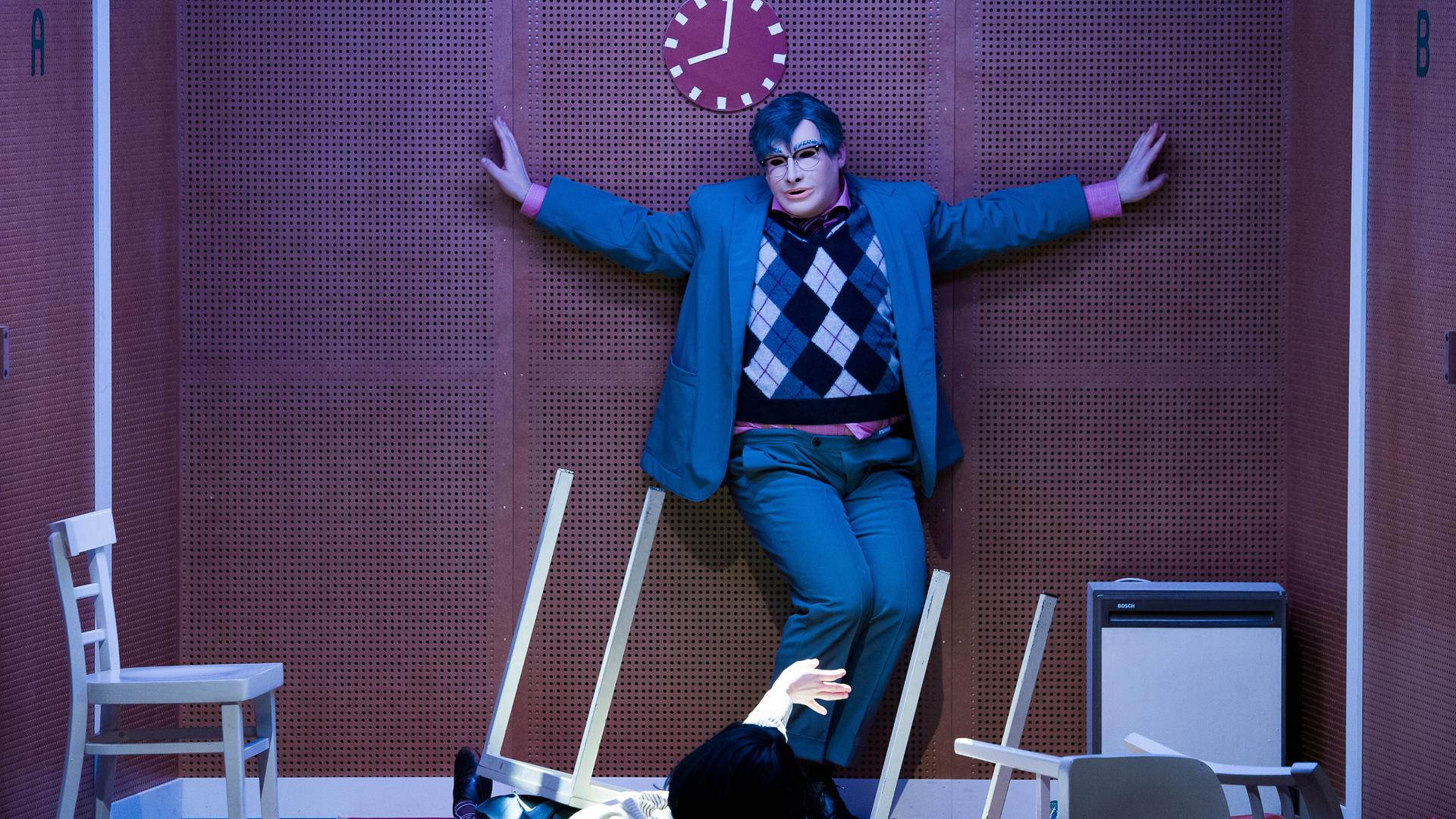 Szene aus dem Theaterstück "humanistää!" am Volkstheater in Wien. Eine Person mit ausgebreiteten Armen und mit Maske steht in einem grauen Anzug mit kariertem Pullunder vor einer braunen Wand. Szene aus dem Theaterstück "humanistää!" am Volkstheater in Wien. Eine Person mit ausgebreiteten Armen und mit Maske steht in einem grauen Anzug mit kariertem Pullunder vor einer braunen Wand.