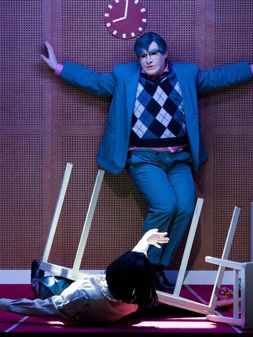 Szene aus dem Theaterstück "humanistää!" am Volkstheater in Wien. Eine Person mit ausgebreiteten Armen und mit Maske steht in einem grauen Anzug mit kariertem Pullunder vor einer braunen Wand.