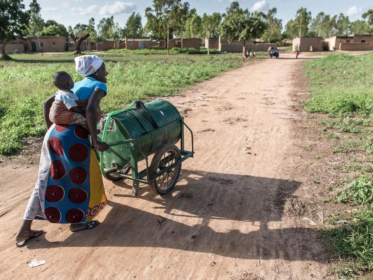 Eine Frau in Ouagadougou / Burkina Faso schiebt einen Trinkwassertank, den sie an einer öffentlichen Wasserzapfstelle befüllt hat, zurück ins Dorf. Man sieht am Horizont das Dorf im Wüstensand. 
