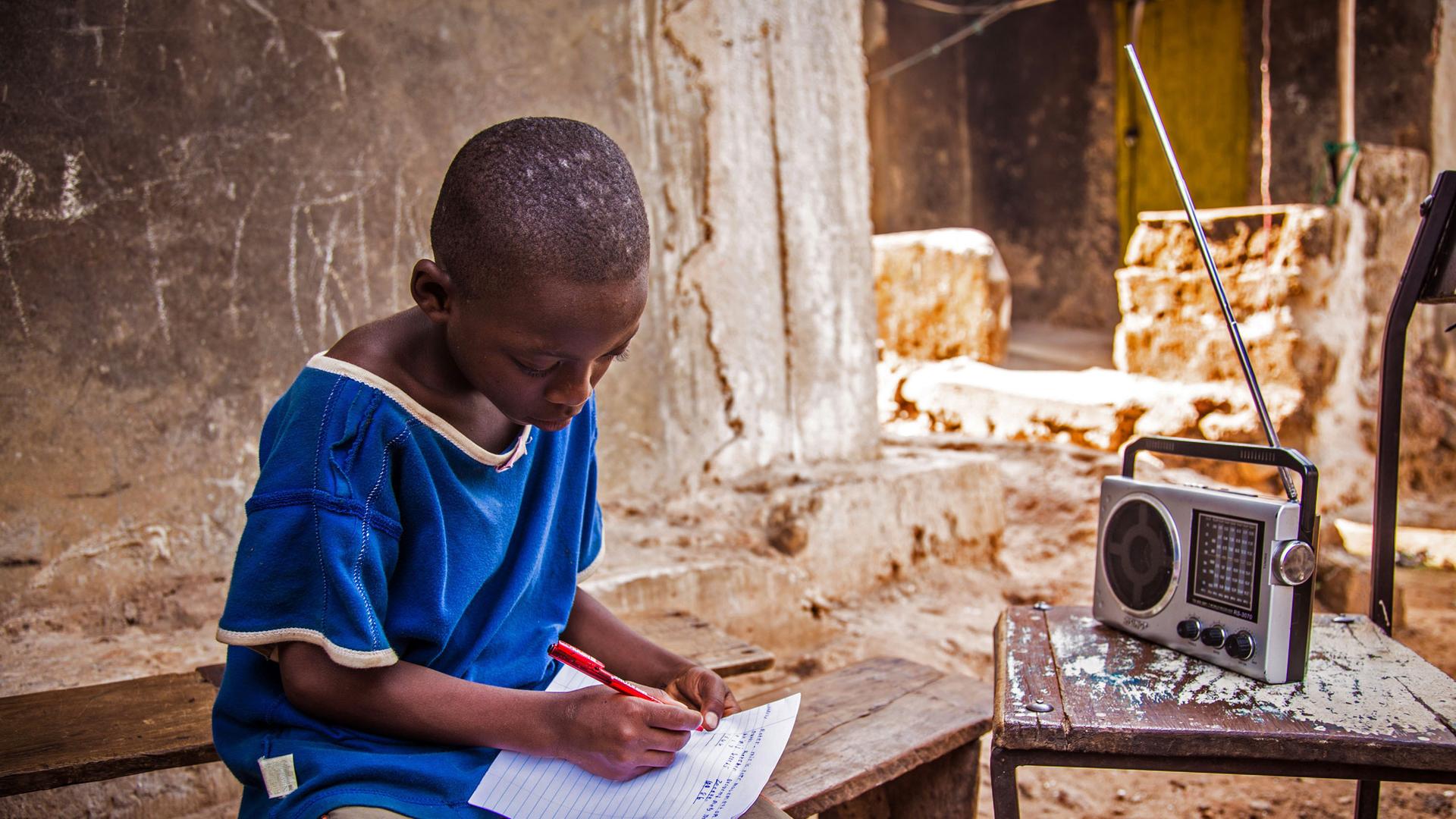 Ein junger sitzt in Sierra Leone mit Stift und Papier vor dem Radiogerät und erledigt seine Schulaufgaben. Ein junger sitzt in Sierra Leone mit Stift und Papier vor dem Radiogerät und erledigt seine Schulaufgaben.