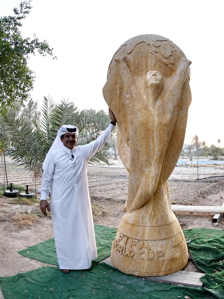 Ein Geschäftsmann in weißem Umhang posiert neben einer beinahe drei Meter hohen Skulptur des FIFA-Pokals aus sandfarbenem Travertin.