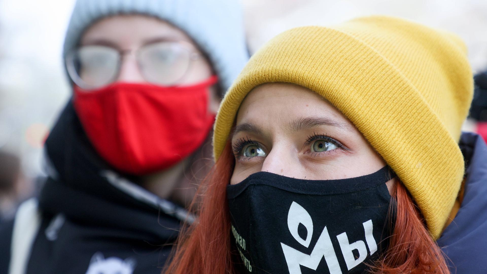Eine junge Frau mit schwarzen Mundschutz und gelber Mütze blickt nach links oben. Auf dem Mundschutz ist das Logo der Organisation Memorial gedruckt. Hinter der Frau steht ein Mann mit rotem Mundschutz. Eine junge Frau mit schwarzen Mundschutz und gelber Mütze blickt nach links oben. Auf dem Mundschutz ist das Logo der Organisation Memorial gedruckt. Hinter der Frau steht ein Mann mit rotem Mundschutz.