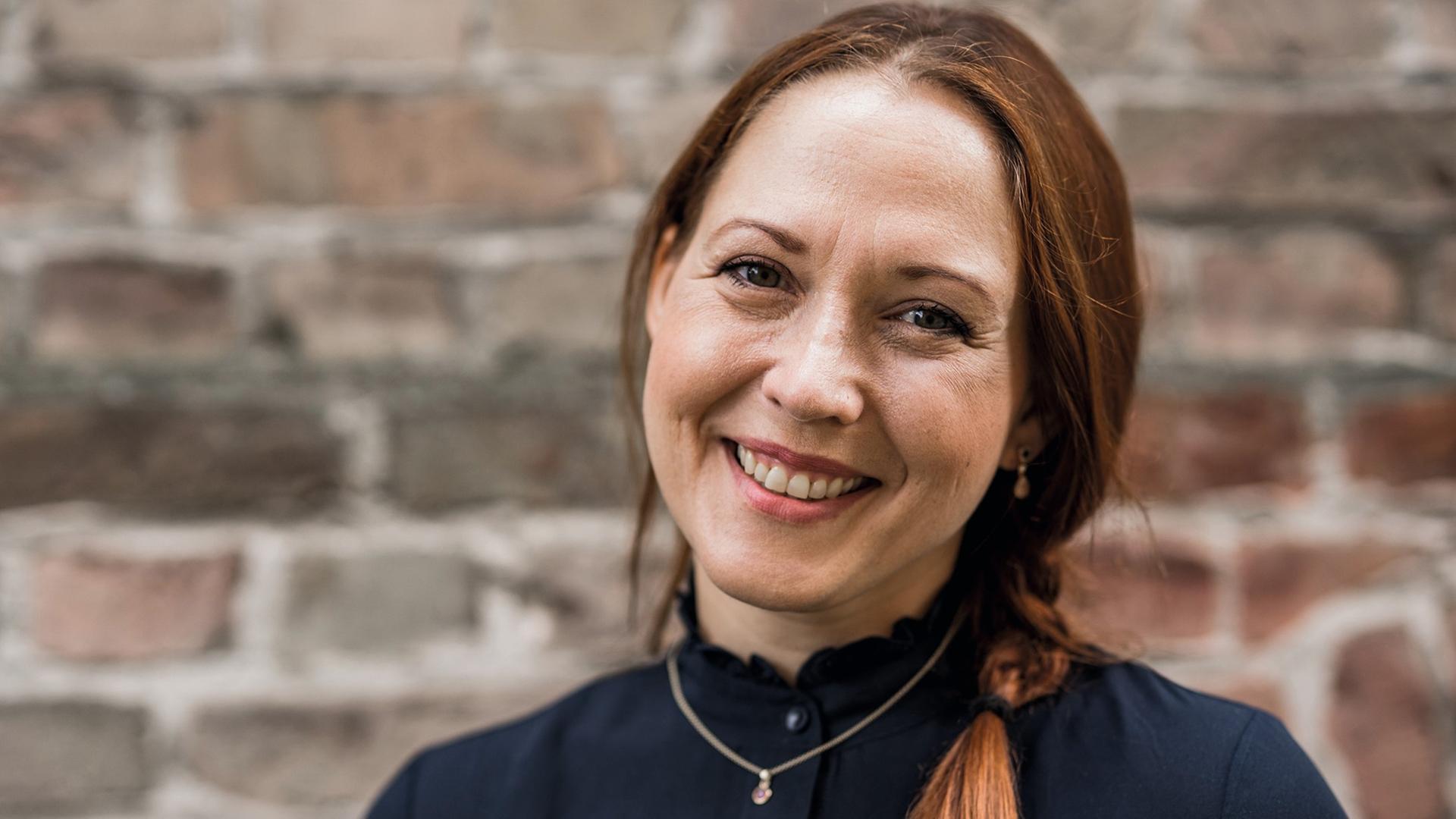 Susanne Mierau schaut in die Kamera. Sie hat lange, rötlich-braune Haare, die sie zu einem Zopf zusammengebunden hat. Sie trägt in dunkles Oberteil und eine goldene Kette mit Anhänger. Im Hingtergrund ist Mauerwerk zu sehen. Susanne Mierau schaut in die Kamera. Sie hat lange, rötlich-braune Haare, die sie zu einem Zopf zusammengebunden hat. Sie trägt in dunkles Oberteil und eine goldene Kette mit Anhänger. Im Hingtergrund ist Mauerwerk zu sehen.