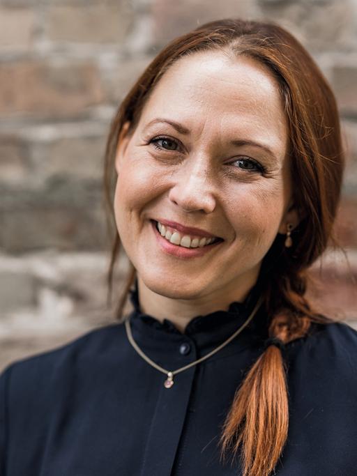 Susanne Mierau schaut in die Kamera. Sie hat lange, rötlich-braune Haare, die sie zu einem Zopf zusammengebunden hat. Sie trägt in dunkles Oberteil und eine goldene Kette mit Anhänger. Im Hingtergrund ist Mauerwerk zu sehen.