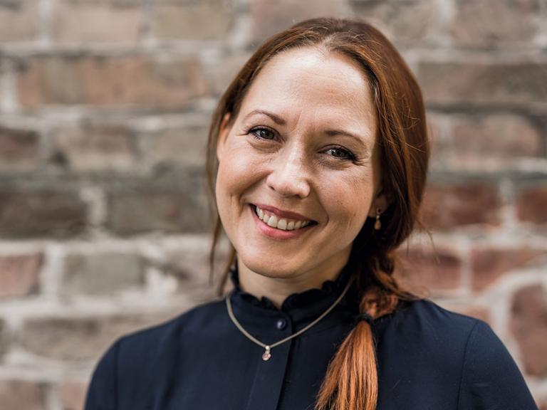 Susanne Mierau schaut in die Kamera. Sie hat lange, rötlich-braune Haare, die sie zu einem Zopf zusammengebunden hat. Sie trägt in dunkles Oberteil und eine goldene Kette mit Anhänger. Im Hingtergrund ist Mauerwerk zu sehen.