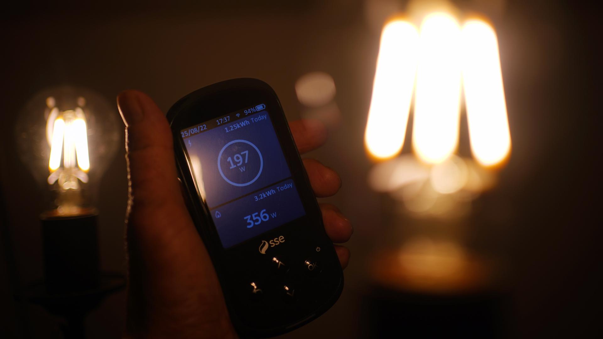 Eine Hand hält einen digitalen Stromzähler in der Hand, im Hintergrund leuchten Glühbirnen