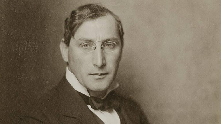 Ein historisches Foto zeigt einen gepflegten Mann mit Seitenscheitel, kleiner Brille und Schal im hohen Kragen.