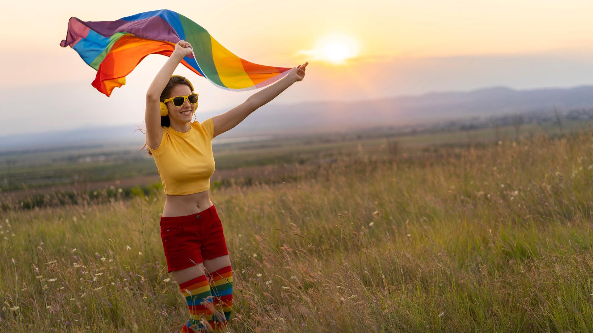 Glückliche junge Frau in sommerlicher bunter Kleidung mit Kopfhörern und Regenbogenfahne vor einem Sonnenuntergang. Glückliche junge Frau in sommerlicher bunter Kleidung mit Kopfhörern und Regenbogenfahne vor einem Sonnenuntergang.