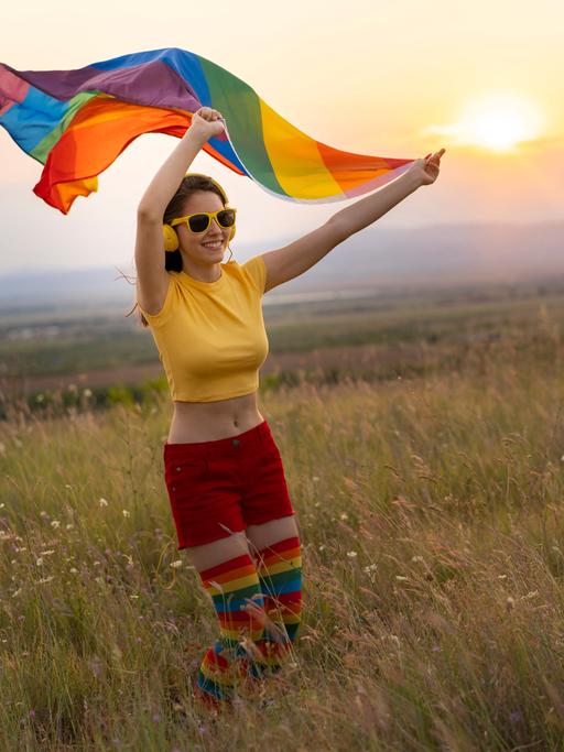 Glückliche junge Frau in sommerlicher bunter Kleidung mit Kopfhörern und Regenbogenfahne vor einem Sonnenuntergang.