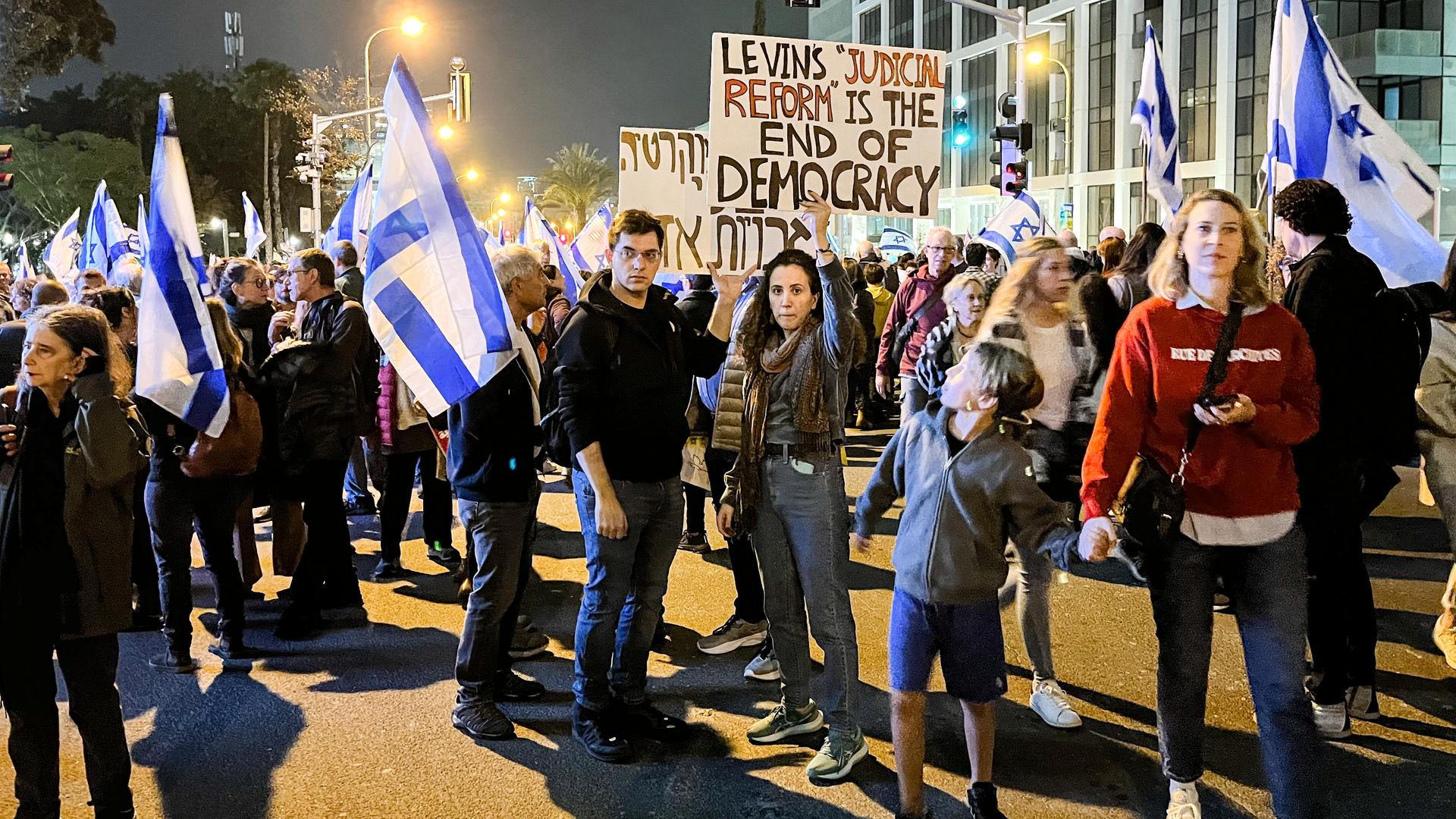 Menschen mit israelischen Fahnen und Protestplakaten gegen die Regierung versammeln sich am Abend auf der Straße.