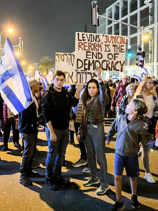 Menschen mit israelischen Fahnen und Protestplakaten gegen die Regierung versammeln sich am Abend auf der Straße.