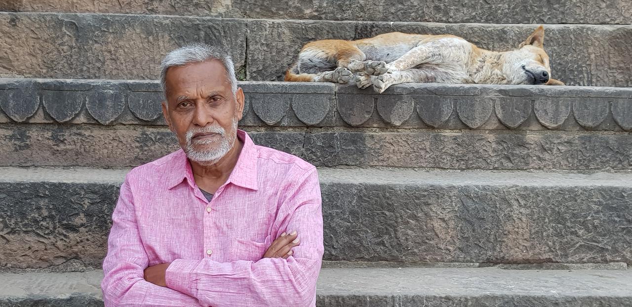 Ein älterer Mann, in einem rosa Hemd und mit verschänkten Armen, steht vor ein Paar Treppenstufen und schaut den Betrachter an. Im Hintergrund ist ein schlafender Hund, der auf den Stufen liegt, zu sehen. Ein älterer Mann, in einem rosa Hemd und mit verschänkten Armen, steht vor ein Paar Treppenstufen und schaut den Betrachter an. Im Hintergrund ist ein schlafender Hund, der auf den Stufen liegt, zu sehen.