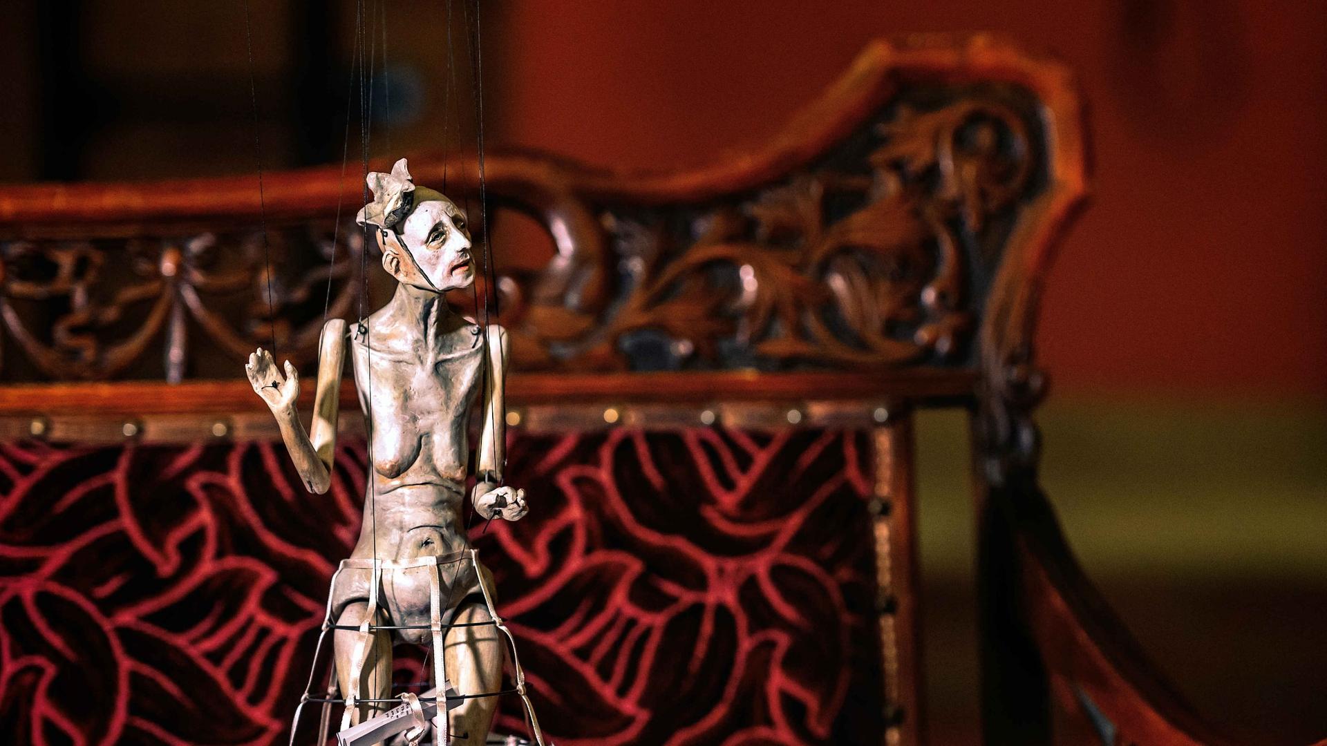 Eine Figur einer alten halbnackten Frau an Marionettenseilen hängend.
