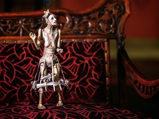 Eine Figur einer alten halbnackten Frau an Marionettenseilen hängend.