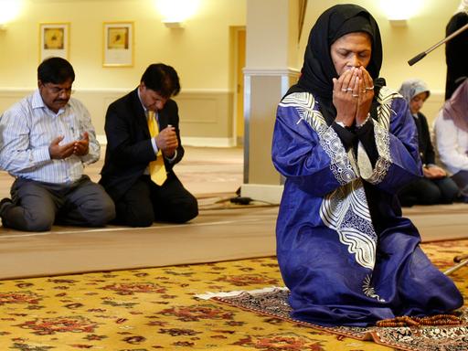 Eine Frau mit schwarzem Kopftuch, in blauem Gewand kniet auf einem Gebetsteppich, hält die Augen geschlossen und die Hände vor das Gesicht. Hinter ihr knien in ähnlicher Haltung Männer und Frauen zum Gebet.