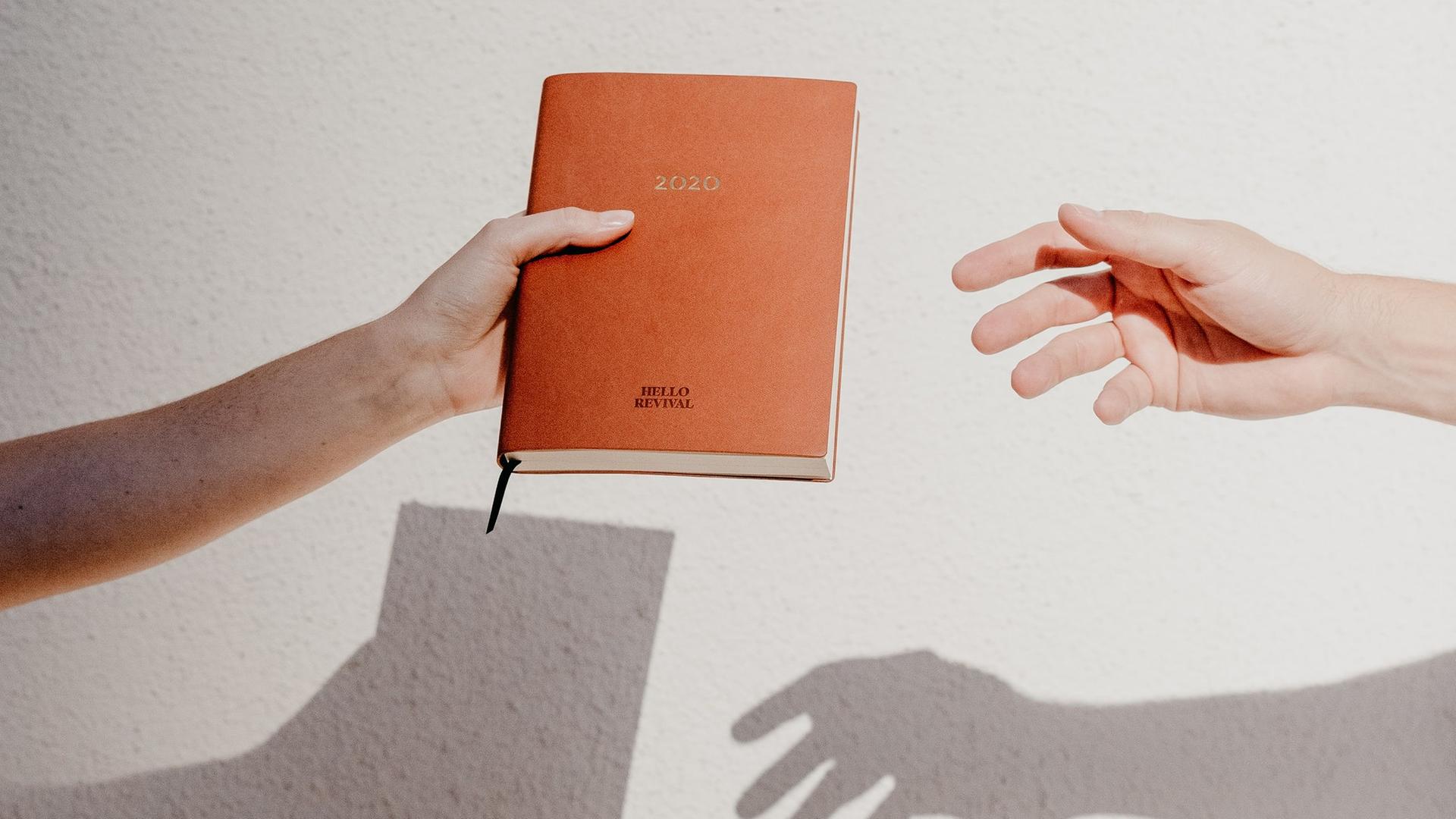 Vor einem weißen Bildhintergrund wird ein in braunes Leder gebundenes Tagebuch von einer Hand in die Nächste weitergereicht. Vor einem weißen Bildhintergrund wird ein in braunes Leder gebundenes Tagebuch von einer Hand in die Nächste weitergereicht.