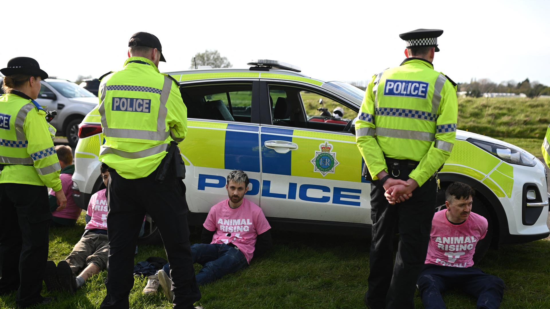 Aktivisten sitzen in Aintree vor einem Polizei-Auto. Polizisten stehen daneben. Aktivisten sitzen in Aintree vor einem Polizei-Auto. Polizisten stehen daneben.