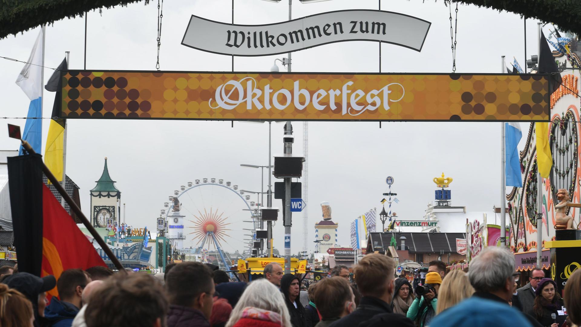 Über den Besuchern hängen zwei Banner mit der Aufschrift "Willkommen zum Oktoberfest". Im Hintergrund ein Riesenrad.